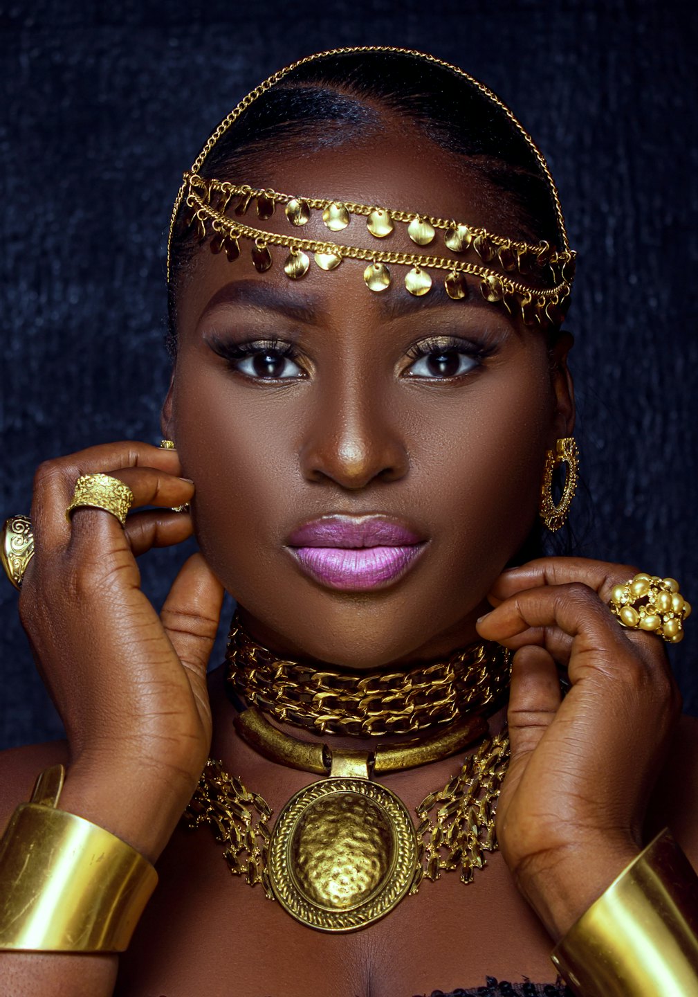 Woman wearing ornate gold jewelry and bold purple lipstick.